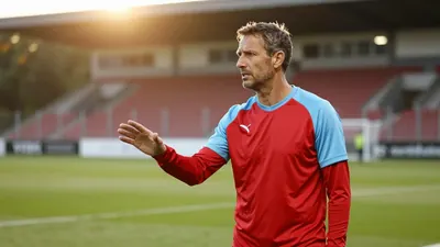 A 35-year-old Annecy coach, wearing a plain red and sky blue polyester training top, stands on the Parc des Sports...